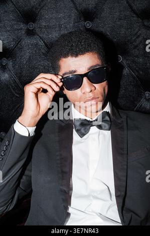 Schwarzer Geschäftsmann mit Anzug und Fliege, der Sonnenbrille verstellt, sitzt im Studio auf einem Stuhl Stockfoto