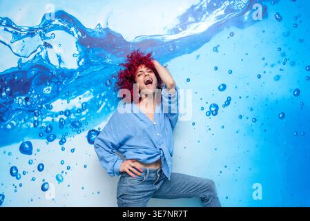 Hispanische Frau mit roten lockigen Haaren lachend mit Hand auf Kopf und Taille stehend mit einem Bein oben vor blauem Wasser spritzen und fallen Wand i... Stockfoto
