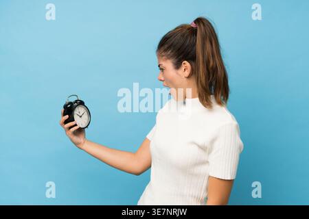 Hispanische Frau mit schockiertem Ausdruck, während sie auf den Wecker vor blauem Hintergrund in stduio blickt Stockfoto