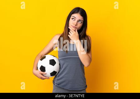Hispanische Frau mit Fußball in der Hand auf dem Kinn, die wegschaut, während sie vor gelbem Hintergrund im Studio steht Stockfoto