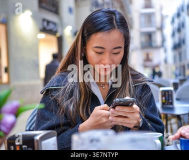 Asiatische Frau mit Smartphone, die tagsüber am Tisch in einem Café im Freien sitzt Stockfoto