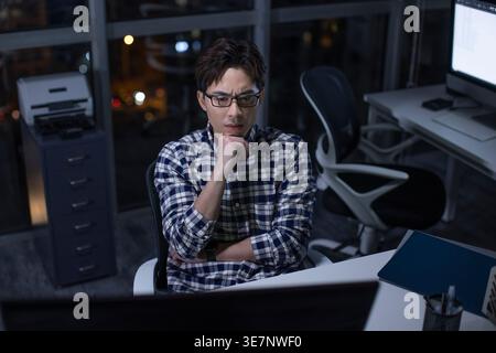 Chinesischer Mann mit Brille in karierten langen Ärmeln, der mit der Hand am Kinn am Tisch sitzt und nachts im Büro auf den Computerbildschirm blickt Stockfoto