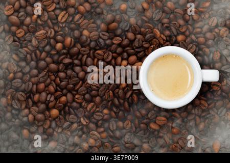 Frisch gebrühter Espresso in einer weißen Tasse, reichhaltige Crema bildet sich oben, umgeben von gerösteten Kaffeebohnen mit aufsteigendem Dampf. Draufsicht Stockfoto