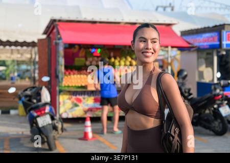 Eine fröhliche, fitte Frau in brauner Aktivkleidung steht auf einem sonnigen Parkplatz in der Nähe eines farbenfrohen tropischen Obststands und Motorrollern, strahlt Wellness und eine positive Stimmung aus Stockfoto