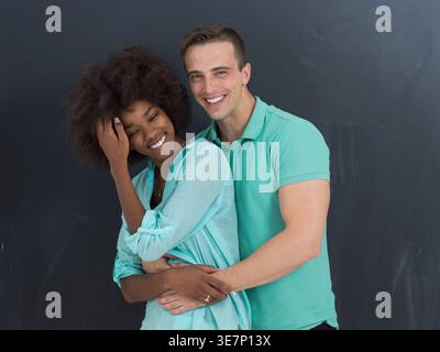 Fröhliche schwarze Frau, die Afrohaar berührt und lacht, mit einem kaukasischen Freund, der sie von hinten an der Tafel im Studio umarmt Stockfoto