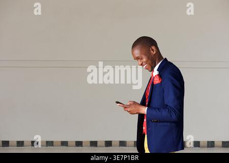 Lächelnder afrikanischer Geschäftsmann mit Smartphone, der tagsüber in der Nähe einer Wand auf der Straße steht Stockfoto