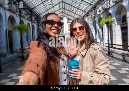 Schwarze und kaukasische Freundinnen tragen Trenchcoat und Sonnenbrille und machen Selfies, während sie tagsüber in der Stadt stehen Stockfoto