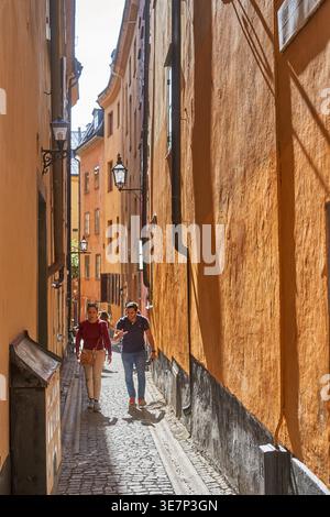 Zwei Personen gehen eine schmale Kopfsteinpflastergasse zwischen hohen, orangefarbenen historischen Gebäuden in der Altstadt von Stockholm hinunter. Die Frau im Vordergrund Stockfoto