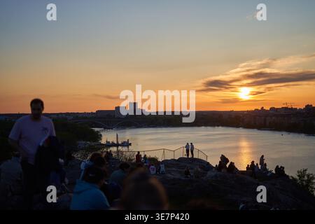 Menschenmenge im Silhouettenstil vor einem lebendigen Sonnenuntergangshimmel, versammelt auf felsigem Gelände mit Blick auf ein Gewässer in Stockholm. Die Sonne geht unter am Stockfoto