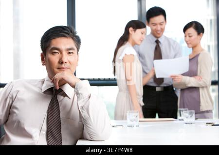 Lächelnder chinesischer Geschäftsmann, der das Kinn auf der Hand beugte, während er am Tisch neben Kollegen sitzt, die im Büro arbeiten Stockfoto