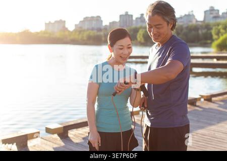 Ein lächelndes chinesisches Paar mittleren Alters, das Smartwatch überprüft, während er am sonnigen Tag in der Nähe des Sees im Park steht Stockfoto