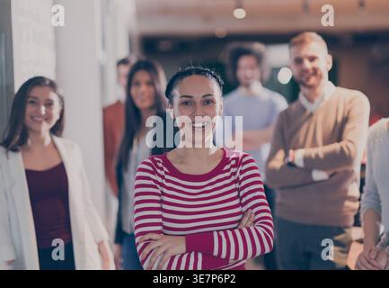 Lächelnde hispanische Geschäftsfrau in gestreiftem Langarmshirt mit überkreuzten Armen und verschiedenen Kollegen im Büro Stockfoto