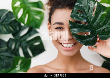 Schwarze Frau, die ein Blatt hält, das ihr Auge bedeckt, während sie im Studio mit großen Blättern an der Wand steht Stockfoto