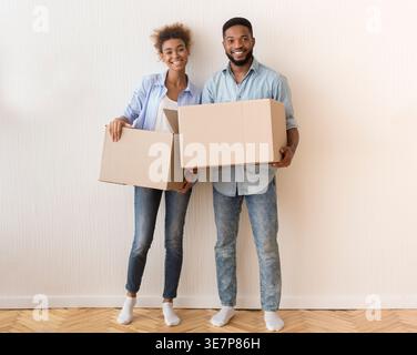 Lächelndes schwarzes Paar hält Umzugskästen, die in einem neuen Zuhause an der weißen Wand stehen Stockfoto