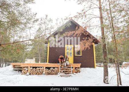 Lächelndes schwarzes Paar in Winterkleidung, das sich ansieht, während es auf der hölzernen Veranda ihres Landhauses sitzt Stockfoto