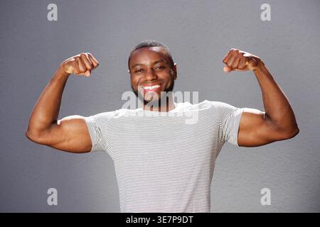 Lächelnder Schwarzer Mann, der bizepsartig vor grauem Hintergrund steht Stockfoto