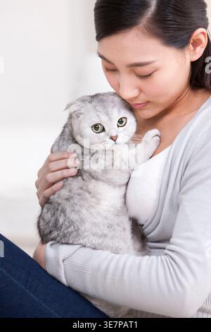 Lächelnde Chinesin, die schottische Faltkatze trägt und umarmt, während sie zu Hause auf dem Sofa sitzt Stockfoto