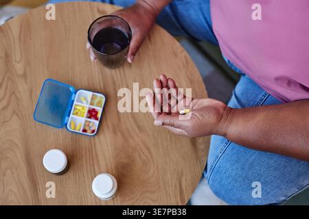 Über dem Blick auf eine schwarze Frau mittleren Alters, die eine Kapsel und ein Glas Wasser hält, während sie zu Hause am Tisch im Wohnzimmer sitzt Stockfoto