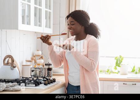 Schwarze Frau kocht und probiert Essen mit Holzlöffel, der zu Hause an der Küchentheke steht Stockfoto