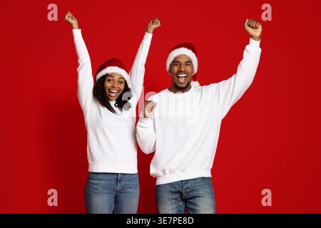 Fröhliches schwarzes Paar mit Weihnachtsmännern, das mit geballten Fäusten vor rotem Hintergrund im Studio steht Stockfoto