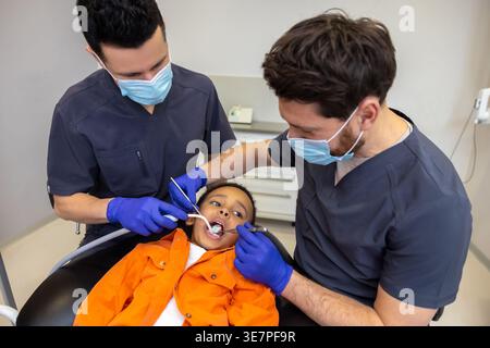 Schwarzer Junge bei zahnärztlicher Behandlung mit kaukasischen männlichen Zahnärzten in der Klinik Stockfoto