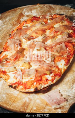 Traditionelle italienische Pinsa romana mit Tomatensauce geschmolzener Mozzarella, pikanten Pilzen und dünnen Scheiben Schinken auf einem bemehlten Holz Stockfoto