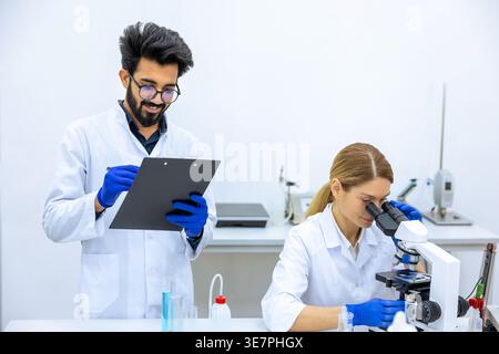 Lächelnder indischer Wissenschaftler, der Labormantel und Latexhandschuhe trägt, macht Notizen, nachdem er einen Beispieltest in der Nähe einer kaukasischen Kollegin mit Mikrosko gemacht hat... Stockfoto