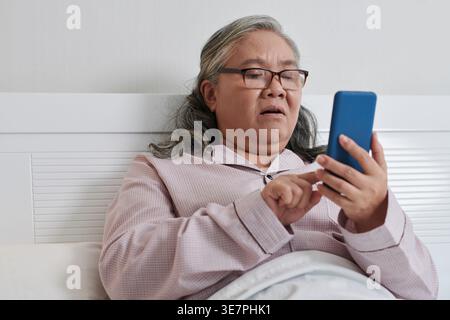 Asiatische ältere Frau mit Brille mit Smartphone, während sie im Bett im Krankenhauszimmer sitzt Stockfoto