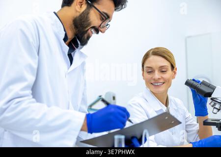 Fröhlicher indischer Wissenschaftler, der Labormantel und Latexhandschuhe trägt, macht Notizen, nachdem er einen Beispieltest in der Nähe von kaukasischen weiblichen Kollegen mit microsc gemacht hat... Stockfoto