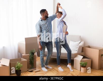 Ein lächelndes schwarzes Paar, das sich ansieht, wie es in einem neuen Zuhause zwischen Umzugskartons tanzt Stockfoto