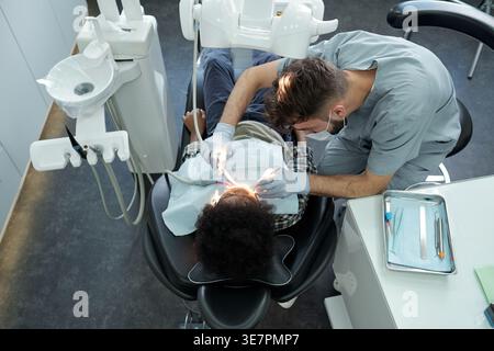 Schwarzer Junge, der mit kaukasischem männlichen Zahnarzt behandelt wird, während er auf einer Zahnarztklinik sitzt Stockfoto