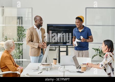 Smiling Black, männliche Programmierer testen neue Virtual-Reality-Brillen, die in der Nähe des Monitors stehen, mit Codes und treffen sich mit weiblichen Kollegen im Büro Stockfoto