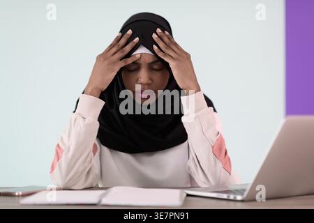 Gestresste schwarze muslimische Geschäftsfrau mit Händen auf dem Kopf und geschlossenen Augen, die im Büro mit Laptop und Notebook am Tisch sitzt Stockfoto