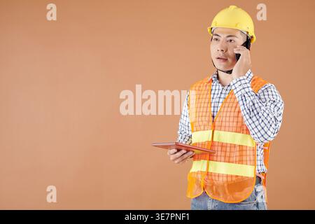 Seriöser asiatischer Bauingenieur, der ein Tablet hält und am Telefon spricht und wegschaut, während er vor orangefarbenem Hintergrund im Studio steht Stockfoto