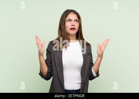 Hispanische Frau mit Blazer, die im Studio mit offenen Armen vor hellgrünem Hintergrund aufblickt Stockfoto