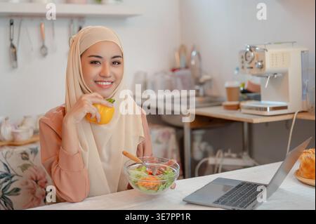 Lächelnde asiatische muslimische Frau im Hijab sitzt am Tisch mit Laptop mit Pfeffer und Salatschüssel in der Küche zu Hause Stockfoto