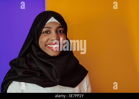Fröhliche schwarze muslimische Frau mit Nasenpiercing, die Hijab trägt und im Studio vor violettem und gelbem Hintergrund steht Stockfoto