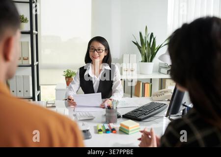 Asiatische Agentin, die Papier hält und Fragen beantwortet, hat sich mit zwei Kunden getroffen, die ihr gegenüber am Tisch im Büro sitzen Stockfoto