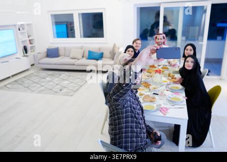 Lächelnde muslimische Familie, die am Tisch sitzt und Selfie auf dem Smartphone macht, bevor sie während des Ramadan-Festmonats zu Hause Iftar zu Abend isst Stockfoto