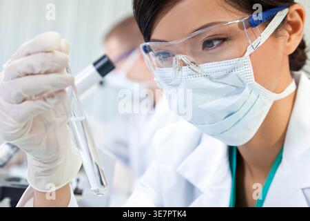 Ernsthafte asiatische Ärztin mit Schutzbrille und Gesichtsmaske beim Blick auf das Reagenzglas im Labor Stockfoto