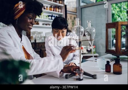 Schwarze Chemikerin öffnet das Ventil des Separationstrichters und lässt es in den Becher fließen, der mit einer japanischen Kollegin in der Vintage-Apotheke steht Stockfoto