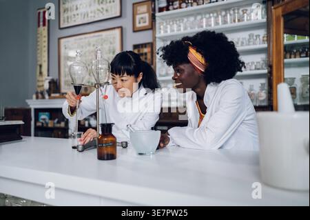 Die konzentrierte japanische Chemikerin und schwarze Kollegin spricht und arbeitet an einem Experiment mit einem Trenntrichter, der in einer Vintage-Apotheke steht Stockfoto