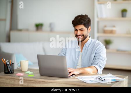 Lächelnder arabischer Mann mit Bart, der am Laptop arbeitet und zu Hause am Schreibtisch sitzt Stockfoto