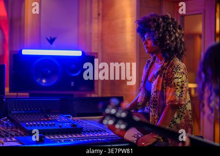 Ernsthafte hispanische Musikerin mit Afro-Blick, die neben einer Mischkonsole mit Neonlicht sitzt und der Aufnahme von Gitarrenspielen zuhört... Stockfoto