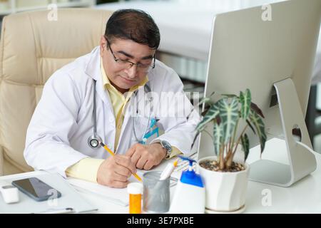 Indischer Arzt mit Stethoskop um den Hals mit Brille am Tisch sitzen und Notizen auf medizinischem Klemmbrett in der Klinik schreiben Stockfoto