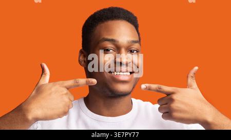 Schwarzer Mann mit weißem Hemd, lächelt und zeigt mit den Fingern auf Zähne und Mund vor orangem Hintergrund Stockfoto