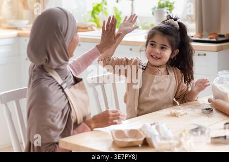 Mutter aus dem Nahen Osten im Hijab und Tochter mit Schürzen, die sich gegenseitig hohe fünf geben, während sie am Tisch mit Zutaten in der Küche zu Hause sitzt Stockfoto