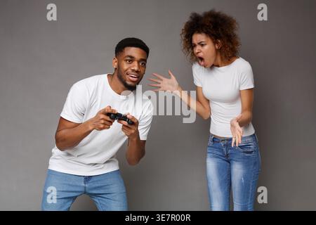 Schwarze Frau brüllt ihren Schwarzen Mann an, während sie im Studio mit Joystick vor grauem Hintergrund spielt Stockfoto