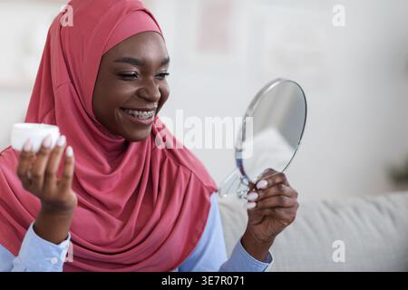Schwarze Frau, die einen Hijab trägt, der ein Glas mit feuchtigkeitsspendender Creme hält und auf den Spiegel blickt, während sie auf dem Sofa zu Hause sitzt Stockfoto
