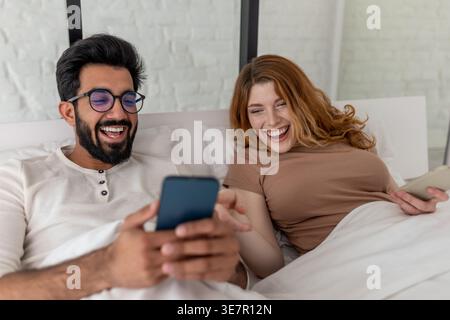 Fröhlicher indischer Ehemann, der eine Brille trägt und der kaukasischen Frau das Handy zeigt, während er sich zu Hause auf dem Bett ausruhte Stockfoto
